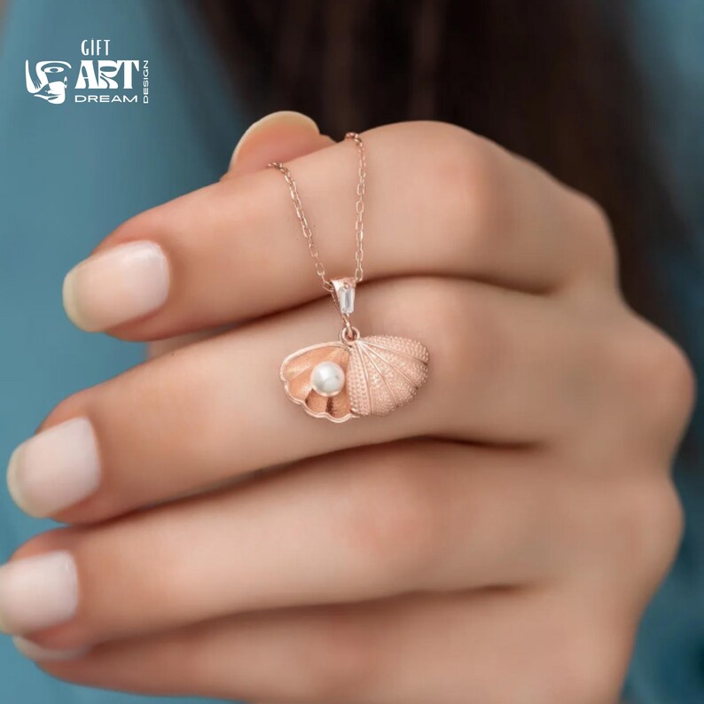 Op de afbeelding: Een ros&eacute;gouden ketting met een schelpenhanger en een parel. De hanger is fijn gedetailleerd en de ketting wordt getoond tegen een zachte achtergrond. De afbeelding bevat ook de tekst "GIFT ART DREAM".