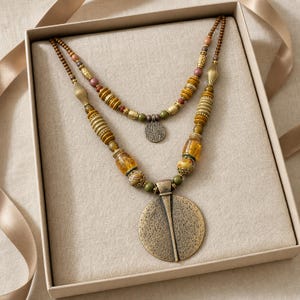 May include: A multi-strand beaded necklace featuring a large, circular, textured bronze-toned pendant. The design incorporates various beads in shades of amber, olive green, brown, and metallic gold, arranged in a layered pattern with smaller circular charms.