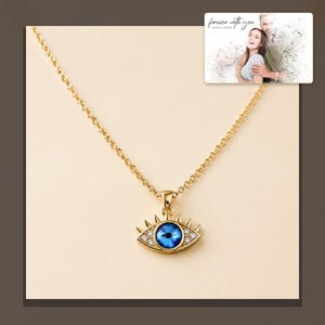 Gold-tone necklace with a blue eye-shaped pendant, alternate framed view. A personalized greeting-card example appears in the corner.