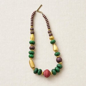 May include: A necklace featuring a sequence of round and elongated beads in shades of purple, green, and red, interspersed with smaller metallic gold-toned spacers. The piece includes a gold-toned metal chain extender with a lobster claw clasp.