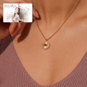 Gold-tone necklace with a round sunburst and black eye detail, worn view. A personalized greeting-card example appears in the corner.