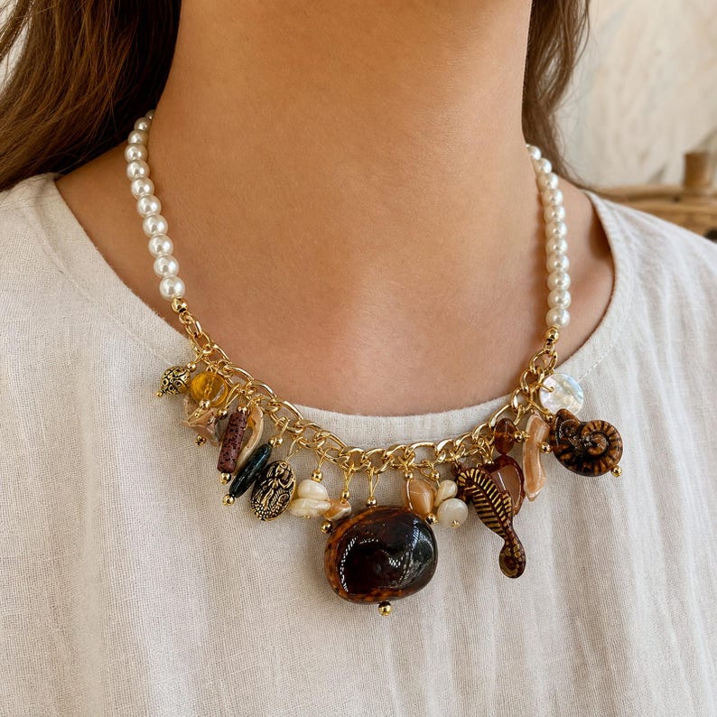 May include: A gold-tone chain necklace featuring a strand of white pearls and a collection of dangling charms, including various brown and tan seashells, polished stones, and a seahorse-shaped pendant, all suspended from a metallic link chain.