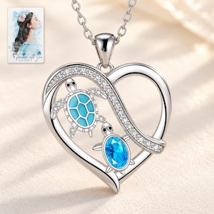 May include: A silver heart-shaped pendant necklace featuring two sea turtles. One turtle has a light blue shell, and the other has a bright blue oval gemstone shell. The heart frame is accented with small clear stones. A small card shows a portrait.