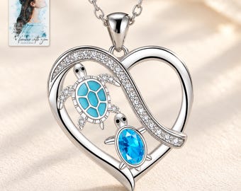May include: A silver heart-shaped pendant necklace featuring two sea turtles. One turtle has a light blue shell, and the other has a bright blue oval gemstone shell. The heart frame is accented with small clear stones. A small card shows a portrait.