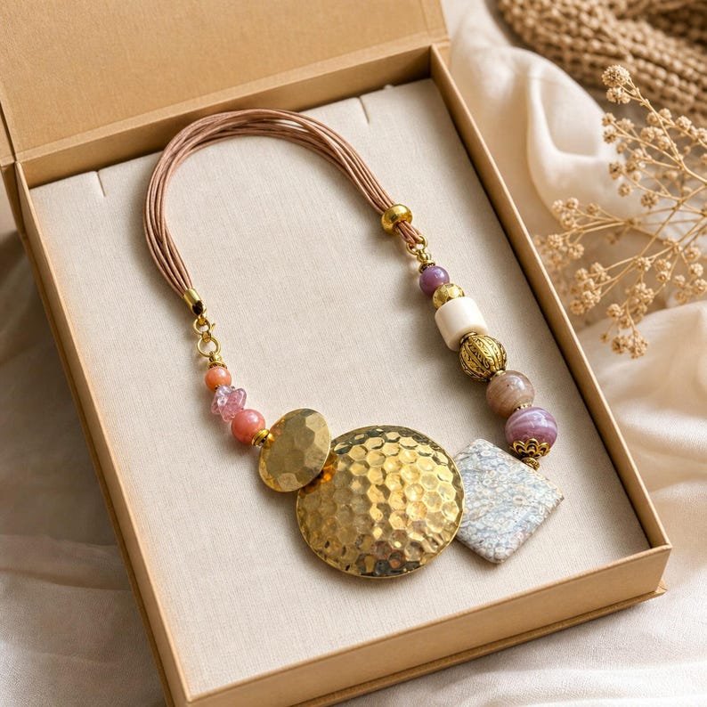 Puede incluir: Un collar de cordones m&uacute;ltiples que presenta un gran colgante circular de tono dorado martillado, una cuenta de piedra cuadrada texturizada y un surtido de cuentas m&aacute;s peque&ntilde;as en tonos rosa, morado, crema y oro met&aacute;lico, dispuestas en un patr&oacute;n.