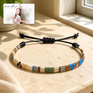 May include: A woven bracelet featuring a geometric pattern in beige, black, green, and blue threads with an adjustable black cord closure. Above the bracelet, a rectangular card displays a photo of a couple with the text "forever with you".