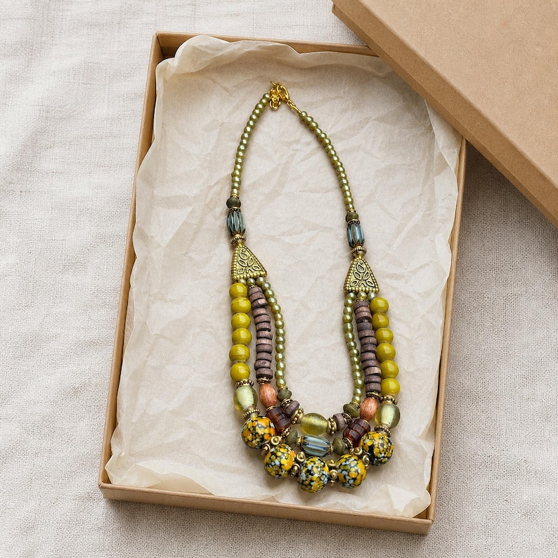 May include: A multi-strand beaded necklace featuring olive green, brown, and blue beads with metallic gold accents. The necklace includes cylindrical, spherical, and patterned floral beads connected by triangular metal findings in a box.