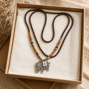 May include: A long necklace featuring dark brown wooden beads and a silver-toned elephant pendant with intricate filigree cutouts. The necklace includes accent beads in shades of mustard yellow, red, black, and green, arranged symmetrically.
