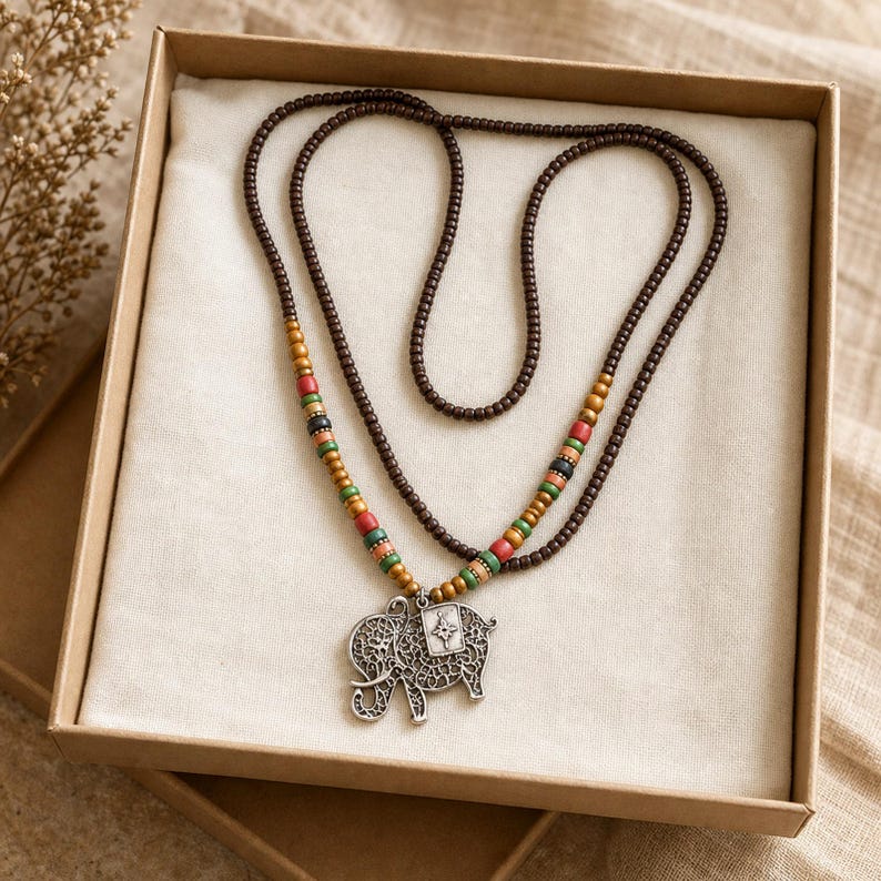 May include: A long necklace featuring dark brown wooden beads and a silver-toned elephant pendant with intricate filigree cutouts. The necklace includes accent beads in shades of mustard yellow, red, black, and green, arranged symmetrically.