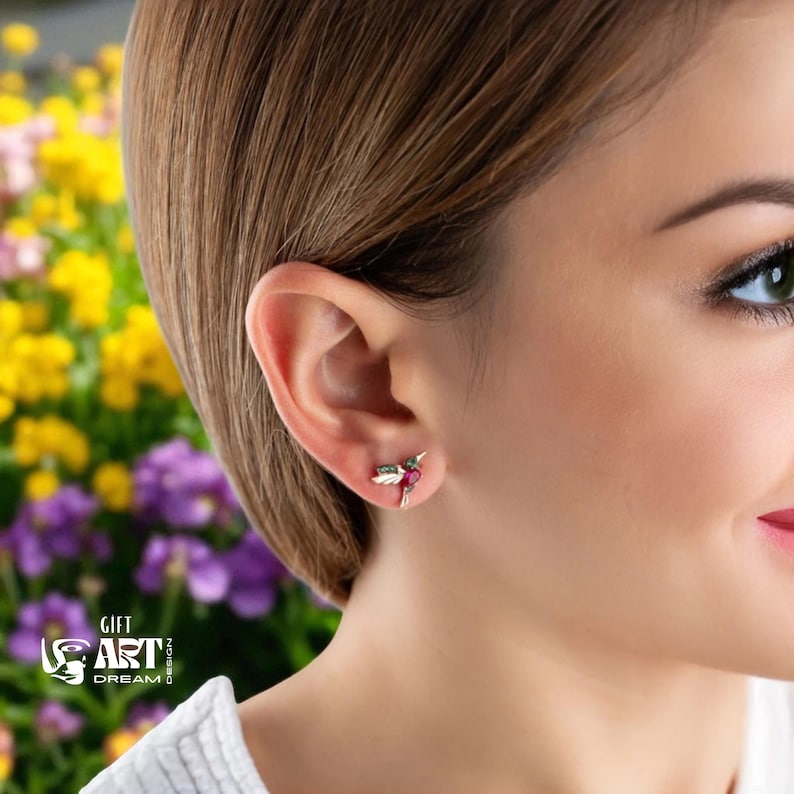 May include: A close-up of a person's ear wearing a small hummingbird earring. The earring features a red gemstone and green accents. The background is blurred, showing yellow and purple flowers. The text "GIFT ART DREAM DESIGN" is visible.