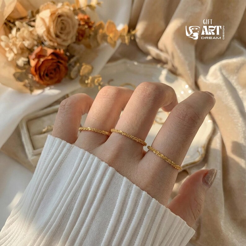 May include: Close-up of several gold-colored rings with a textured surface, worn on fingers. The rings are thin and delicate, reflecting light. The background includes a white sweater, a bouquet of dried flowers, and a logo with the words "GIFT ART DREAM DESIGN".