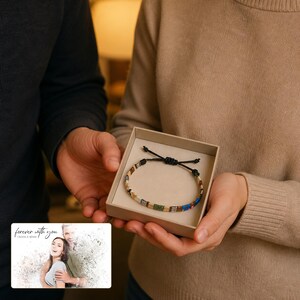 May include: A beaded bracelet with a black cord closure, featuring a pattern of tan, green, white, and blue beads, presented in a square beige gift box. A small card displays the text "forever with you" above the names "OLIVIA & ADAM".