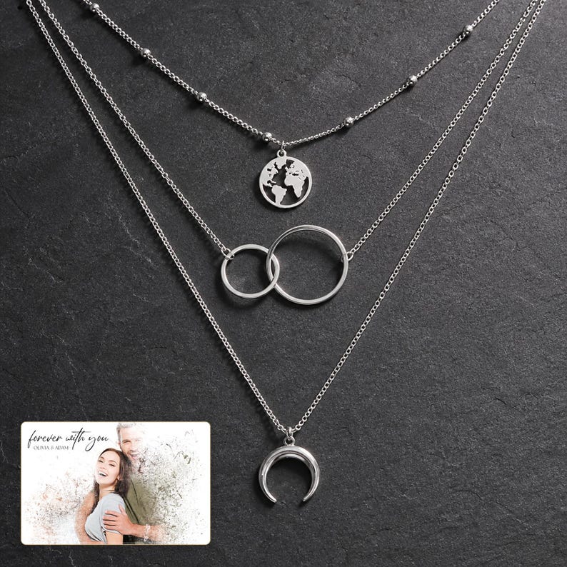 May include: A layered silver-tone necklace featuring three distinct chains. The top chain has small metal beads and a circular world map pendant. The middle chain displays two interlocking rings. The bottom chain features a crescent moon pendant.