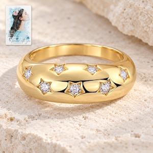 May include: A polished gold band ring featuring a wide, domed profile with six embedded star-shaped settings, each containing a small, clear, round-cut gemstone. The ring is displayed alongside a small rectangular card with a watercolor illustration.