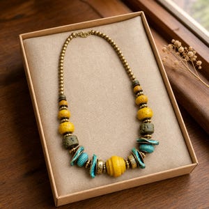 Puede incluir: Un collar de cuentas con una mezcla de cuentas texturizadas redondas en amarillo, verde oliva y azul turquesa, intercaladas con peque&ntilde;as cuentas espaciadoras doradas y un cierre dorado, dispuesto en c&iacute;rculo sobre una base beige.