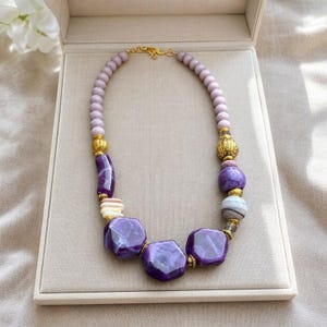 May include: A beaded necklace featuring a mix of light purple round beads, larger faceted deep purple geometric beads, and textured gold-tone metal spacers. The necklace is secured with a gold-tone metal hook clasp at the top center.