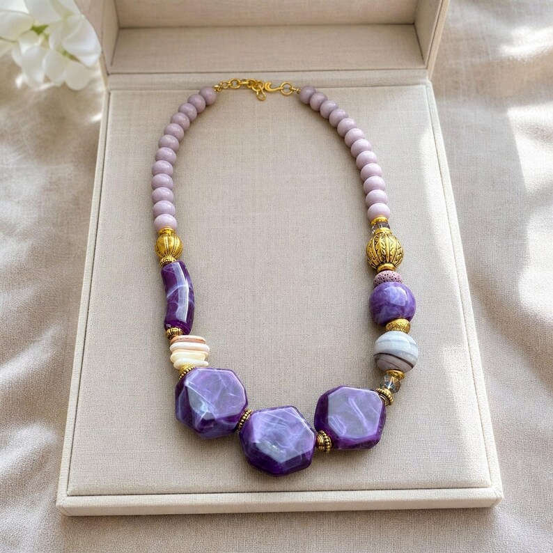 May include: A beaded necklace featuring a mix of light purple round beads, larger faceted deep purple geometric beads, and textured gold-tone metal spacers. The necklace is secured with a gold-tone metal hook clasp at the top center.