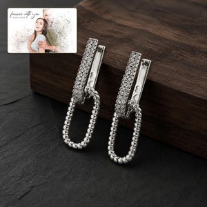 May include: A pair of silver-toned earrings featuring a rectangular top bar encrusted with small clear stones and a dangling oval link with a beaded texture. The earrings are displayed alongside a small rectangular card with the text forever with you.