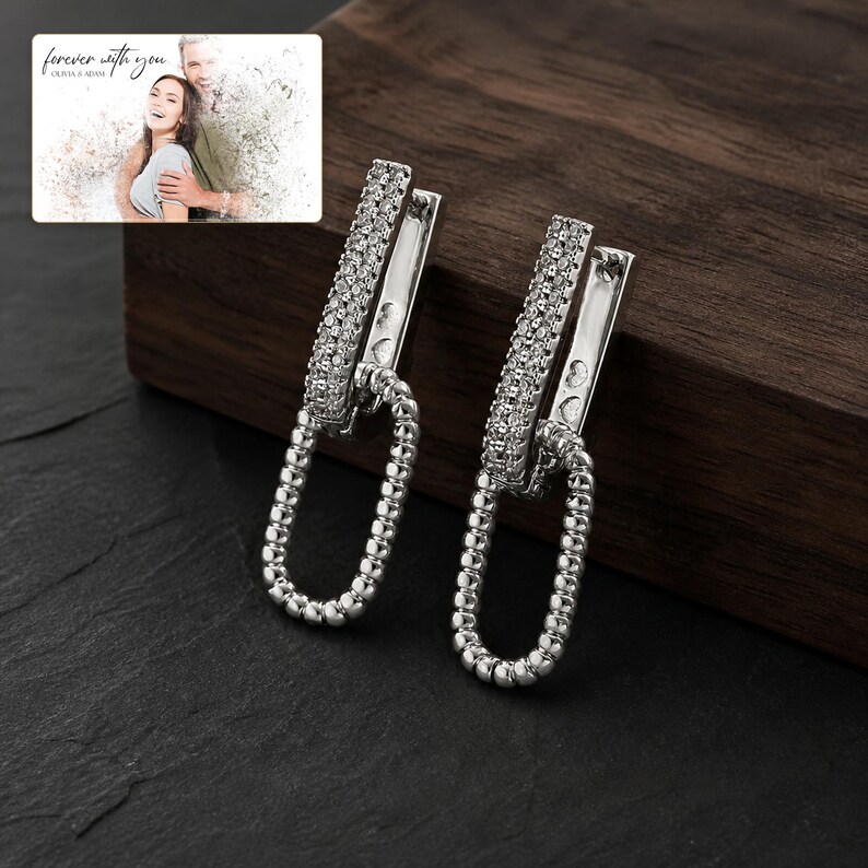May include: A pair of silver-toned earrings featuring a rectangular top bar encrusted with small clear stones and a dangling oval link with a beaded texture. The earrings are displayed alongside a small rectangular card with the text forever with you.