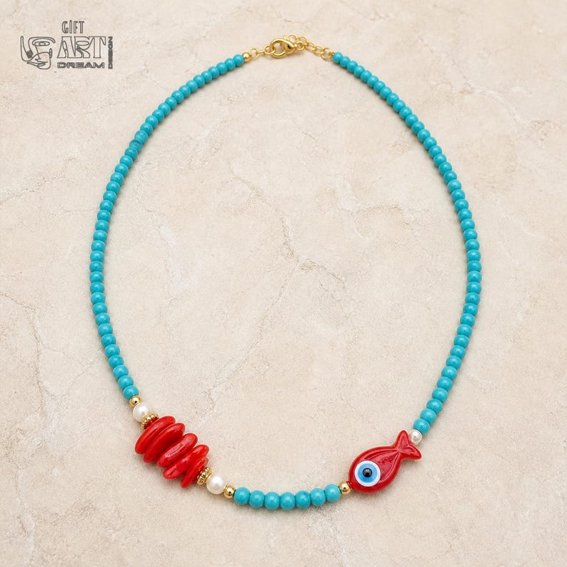 May include: A beaded necklace featuring small turquoise-colored beads, a cluster of four red oval beads, a single red fish-shaped bead with a blue and white evil eye center, small white pearls, and gold-toned metal spacers with a gold-toned clasp.