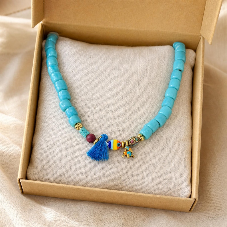 May include: A beaded necklace featuring cylindrical turquoise-colored beads, a small blue tassel, a multi-colored striped bead, a dark red bead, and gold-toned metal accents, presented inside a square cardboard box on a light fabric cushion.