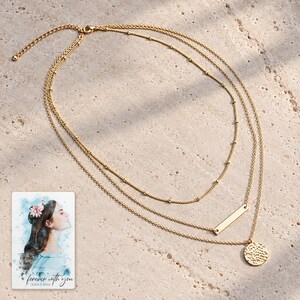 Complete gold-tone three-layer necklace laid large on pale travertine, showing the shared clasp, extender, satellite chain, slim bar and hammered coin. A personalized digital greeting card is shown alongside.