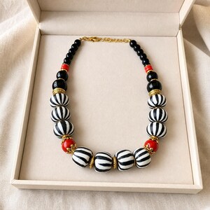 May include: A beaded necklace featuring a sequence of black, white zebra-striped, and bright red spherical beads. The beads are accented with small gold-toned metal spacers and caps, connected by a gold-toned metal chain with a lobster claw clasp.