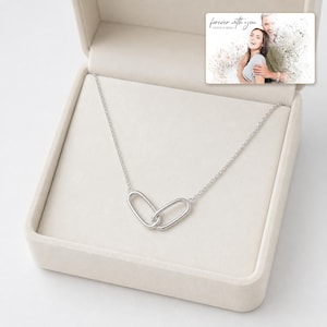 May include: A silver-toned necklace featuring two interlocking oval links as a pendant, presented inside a beige jewelry box. A rectangular card is tucked into the lid, displaying a photo of a couple and the text "forever with you OLIVIA & ADAM."