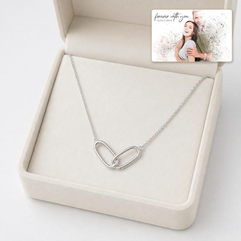 May include: A silver-toned necklace featuring two interlocking oval links as a pendant, presented inside a beige jewelry box. A rectangular card is tucked into the lid, displaying a photo of a couple and the text "forever with you OLIVIA & ADAM."