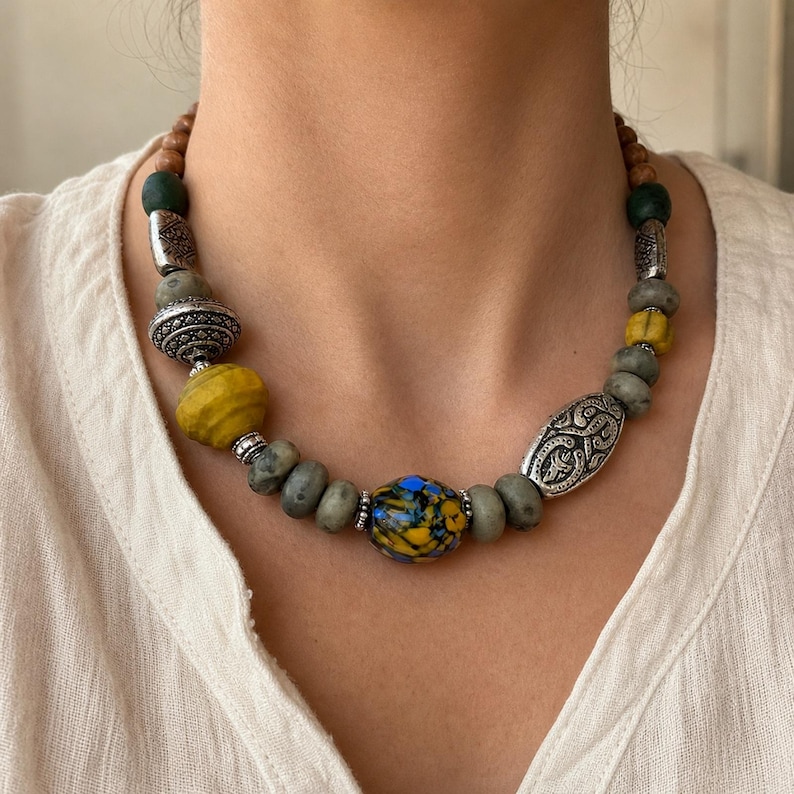 Puede incluir: Un collar de cuentas que presenta una mezcla de cuentas redondas de piedra gris, cuentas amarillas texturizadas y una cuenta central con patrones jaspeados en azul y amarillo. Incluye espaciadores de metal plateado con detalles en relieve.