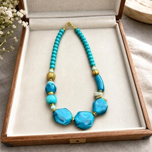 May include: A necklace featuring a strand of turquoise-colored beads in varying shapes and sizes, interspersed with metallic gold-toned spherical spacers. The design includes smooth round beads, larger faceted blue pieces, and small gold accents.