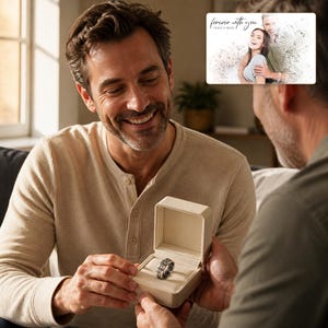May include: A silver-toned metal ring with an intricate, textured pattern sits inside a small, open, cream-coloured rectangular jewellery box. The ring features a repeating design around its entire band, presented in a clean, neutral-coloured case.