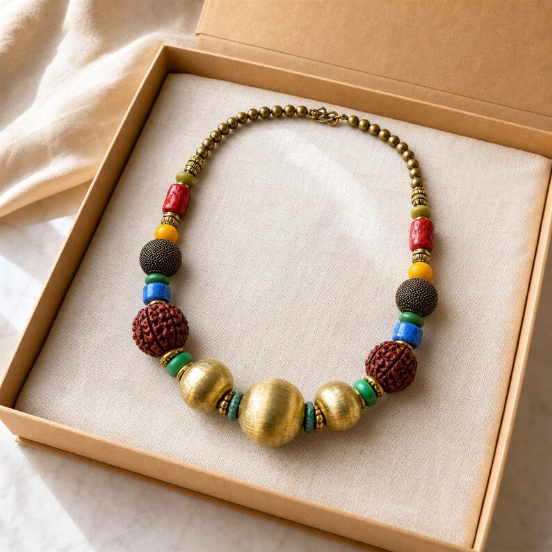 Op de afbeelding: Een ketting met een verscheidenheid aan kralen, waaronder grote geborstelde goudkleurige bollen, getextureerde donkerbruine bollen, gladde rode cilinders, gele bollen, blauwe cilinders, groene tussenstukjes en kleine metalen goudkleurige kralen op draad.