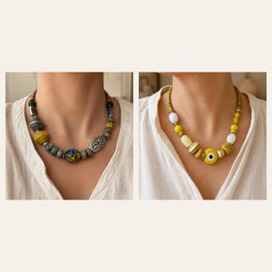 Puede incluir: Dos collares de cuentas. El collar de la izquierda presenta cuentas grises, amarillas y plateadas con una cuenta central de patr&oacute;n multicolor. El collar de la derecha presenta cuentas amarillas, blancas y doradas, incluyendo una cuenta de ojo.