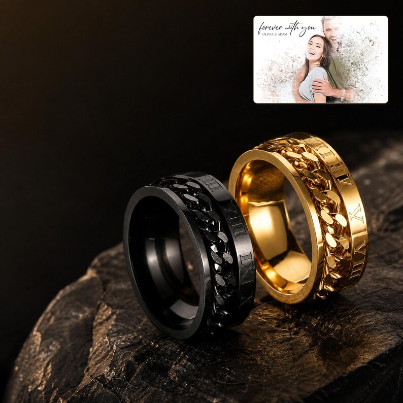 May include: Two spinner rings, one matte black and one polished gold, featuring a central rotating chain link band and engraved Roman numerals. The rings are displayed alongside a small rectangular card with the text "forever with you" and "OLIVIA & ADAM" above a faded photograph of a couple.