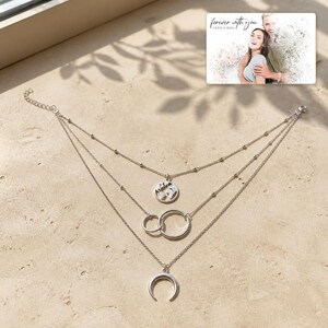 May include: A silver-toned multi-strand necklace featuring three distinct layers. The top layer displays a circular world map charm, the middle layer features two interlocking rings, and the bottom layer holds a crescent moon pendant.
