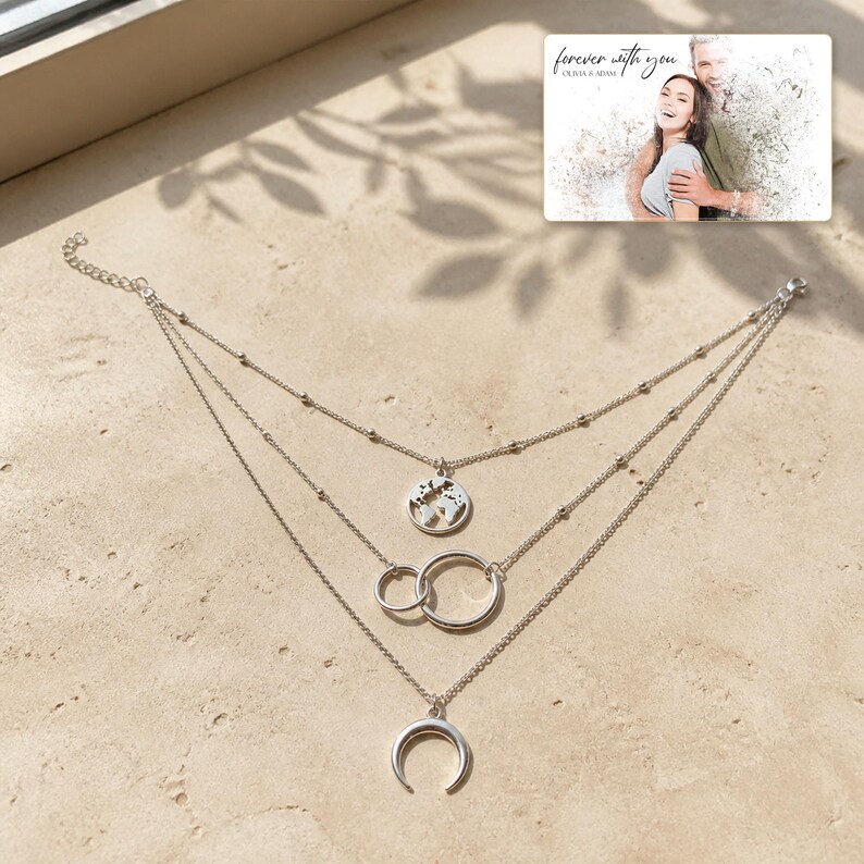 May include: A silver-toned multi-strand necklace featuring three distinct layers. The top layer displays a circular world map charm, the middle layer features two interlocking rings, and the bottom layer holds a crescent moon pendant.