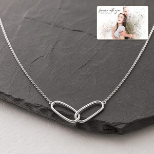 May include: A silver-toned necklace featuring two interlocking oval links as a central pendant. The chain is delicate and metallic. An inset rectangular graphic shows a couple with the text "forever with you" and "OLIVIA & ADAM".