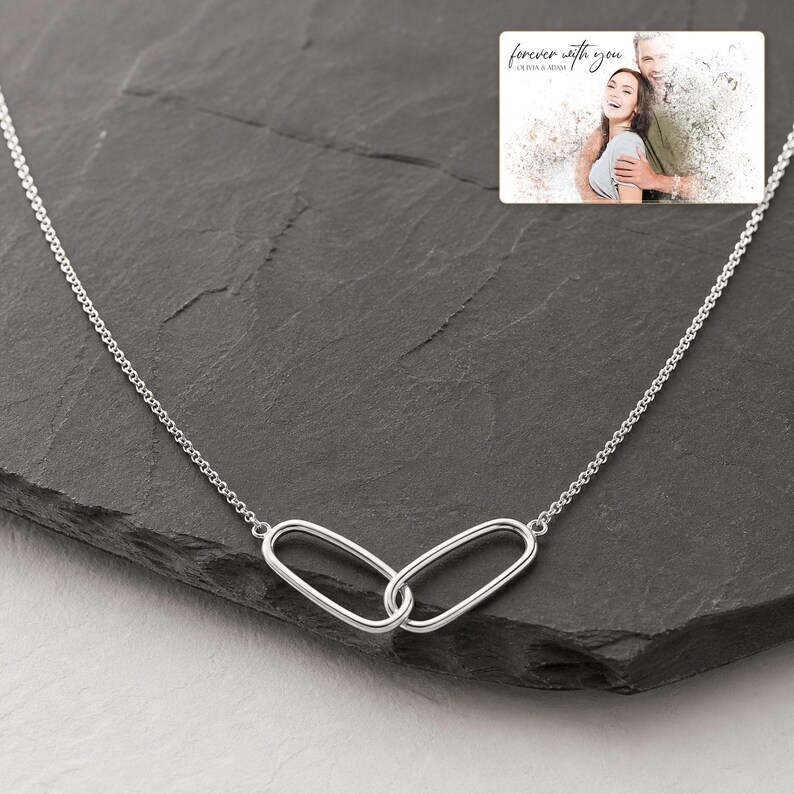 May include: A silver-toned necklace featuring two interlocking oval links as a central pendant. The chain is delicate and metallic. An inset rectangular graphic shows a couple with the text "forever with you" and "OLIVIA & ADAM".