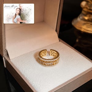 Gold-tone open ring with rows of clear accents, presentation in an open jewelry box. A personalized greeting-card example appears in the corner.