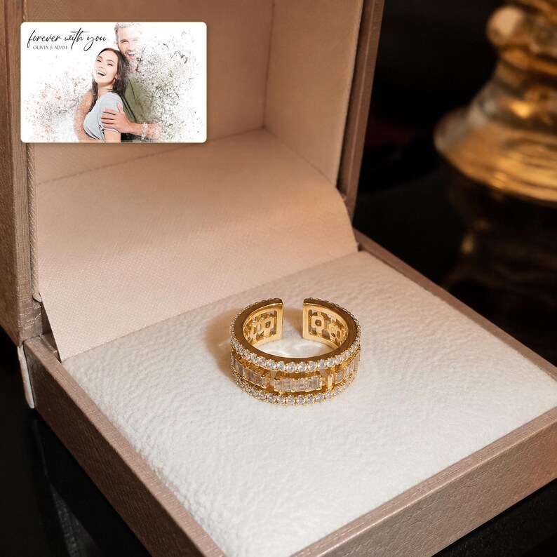 Gold-tone open ring with rows of clear accents, presentation in an open jewelry box. A personalized greeting-card example appears in the corner.