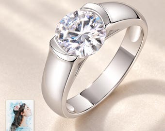 Può includere: Un anello color argento con una grande pietra trasparente rotonda e sfaccettata montata su una fascia a tensione. Una piccola carta rettangolare mostra l'illustrazione di una persona e il testo "forever with you".