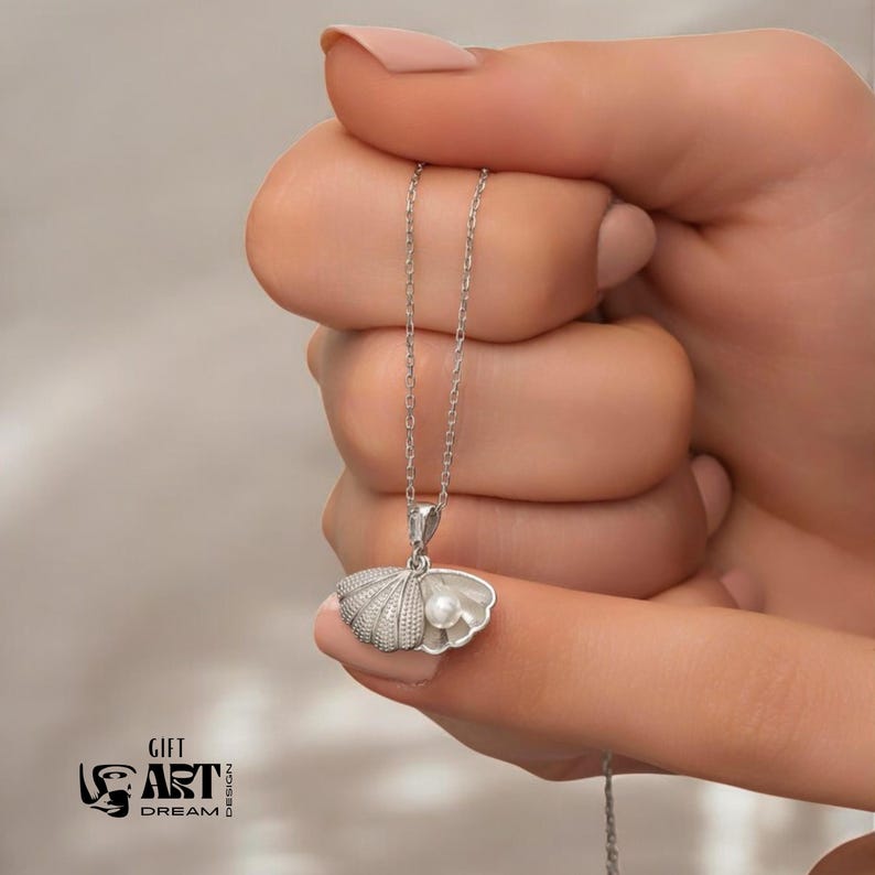 Op de afbeelding: Een zilveren ketting met een schelpenhanger en een parel. De hanger wordt door een hand vastgehouden. De ketting heeft een fijne ketting. De afbeelding bevat ook de tekst "GIFT ART DREAM DESIGN".