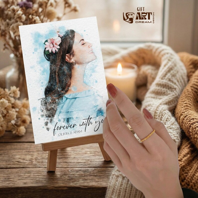 May include: A watercolor print on a small easel, featuring a woman with a flower in her hair, the text "forever with you" and the names Olivia & Adam. A gold ring is on a finger. A lit candle and a cozy sweater are in the background.