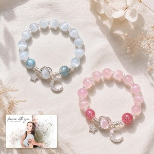 Puede incluir: Dos pulseras de cuentas con perlas ojo de gato en azul claro y rosa, espaciadores plateados y dijes en forma de estrella, luna creciente y planeta. Una peque&ntilde;a tarjeta rectangular muestra el texto "forever with you OLIVA & ADAM".