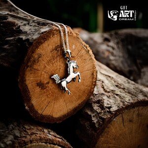 Puede incluir: Collar con colgante de caballo plateado. El colgante presenta un dise&ntilde;o detallado de un caballo encabritado con cuerpo blanco y detalles negros. El collar se muestra sobre una superficie de madera r&uacute;stica. Se ve el texto "GIFT ART DREAM DESIGN".