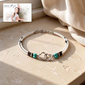 May include: A double-strand bracelet featuring white seed beads, dark brown beads, and turquoise-coloured accents, centred with two interlocking silver-toned heart charms. A small rectangular card displays a photo of a couple with the text "forever with you" and "OLIVIA & ADAM".