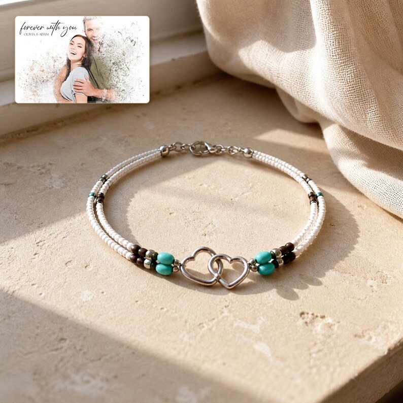 May include: A double-strand bracelet featuring white seed beads, dark brown beads, and turquoise-coloured accents, centred with two interlocking silver-toned heart charms. A small rectangular card displays a photo of a couple with the text "forever with you" and "OLIVIA & ADAM".