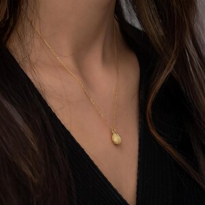 Op de afbeelding: Een goudkleurige ketting met een getextureerde hanger. De hanger heeft een druppelvormig ontwerp. De ketting wordt gedragen tegen een donkere achtergrond.