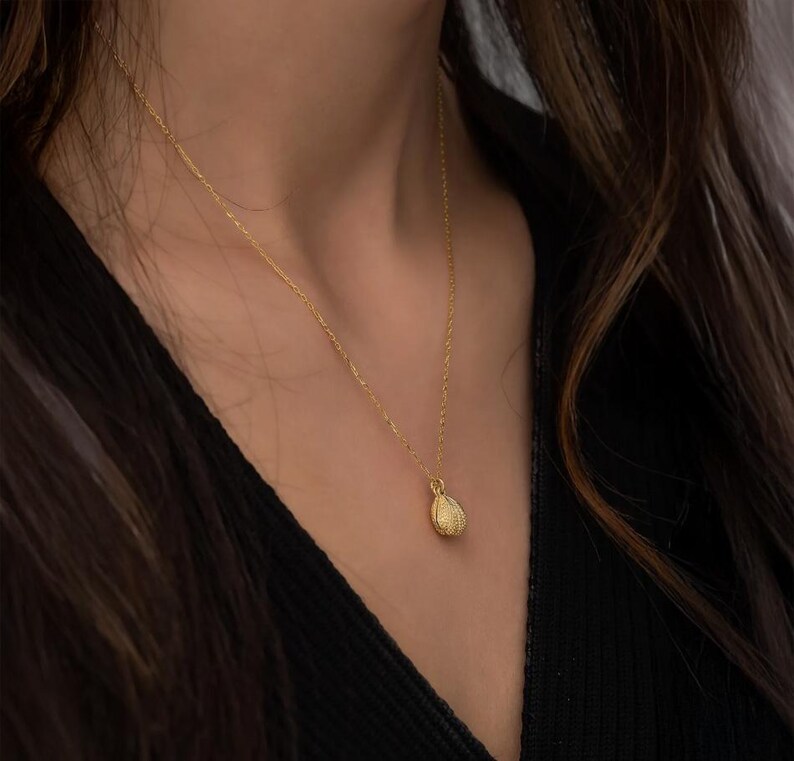 Op de afbeelding: Een goudkleurige ketting met een getextureerde hanger. De hanger heeft een druppelvormig ontwerp. De ketting wordt gedragen tegen een donkere achtergrond.